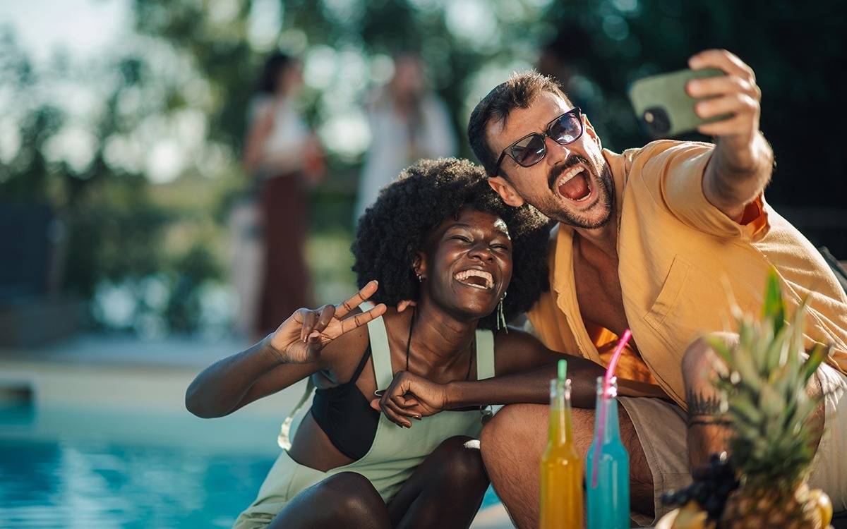 man and woman smiling, taking selfie in front of swimming pool