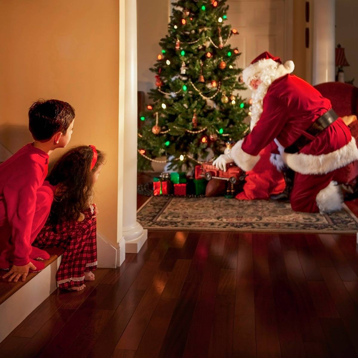 Two children peek on Santa as he leaves presents under their Christmas tree