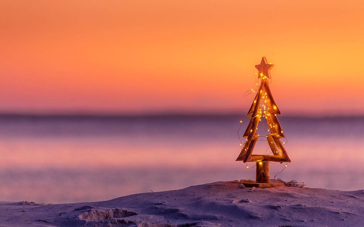 tiny wooden Christmas tree with fairy lights on beach against soft lavender and orange sunset