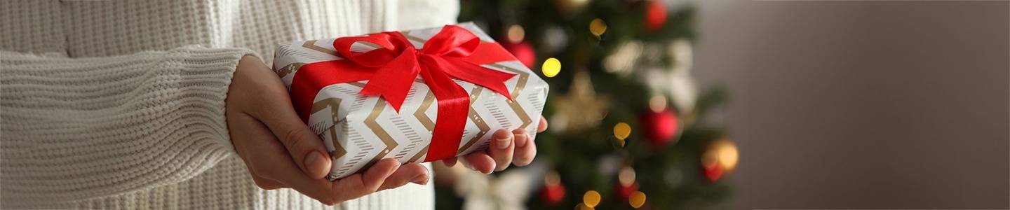 woman wearing white sweater holding wrapped Christmas present with Christmas tree in background