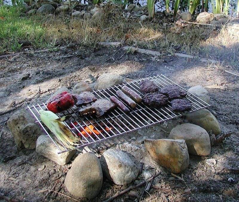 Steel grilling grate over a campfire in a ring of stones, cooking burgers, steak, hot dogs, corn, and pepper