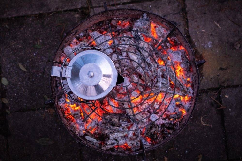 campfire with glowing red coals under a steel grilling grate with metal coffee pot on top
